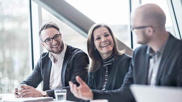 To menn og en kvinne som smiler til hverandre sittende på et møterom